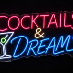 Martini glass icon with Cocktails & Dreams text in pink, white and blue neon on clear acrylic, mounted on a dark wall