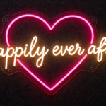 Happily Ever After neon sign in warm white script inside a pink heart outline on clear acrylic, mounted on a dark wall